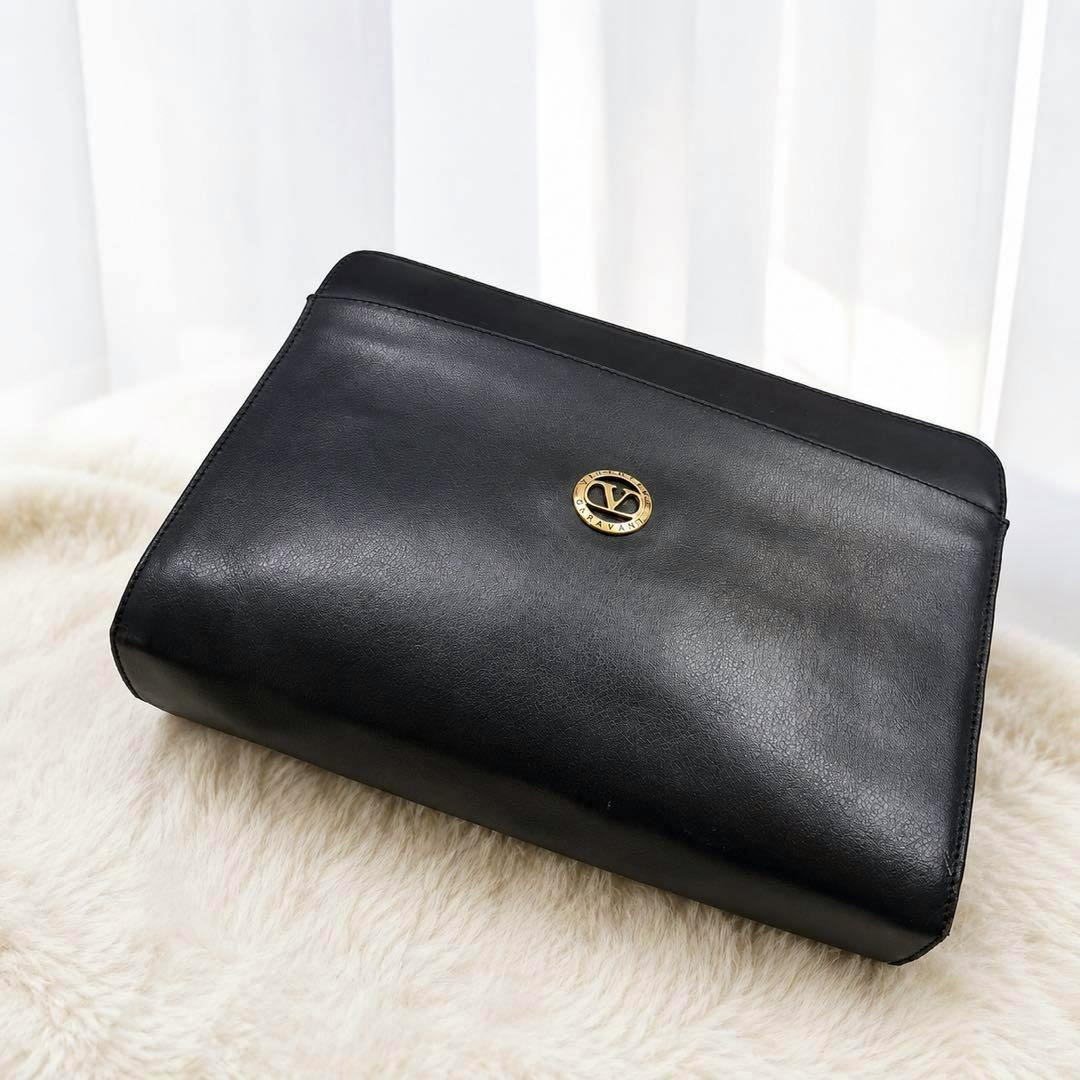 Black Valentino Garavani leather clutch bag with a round gold V logo photographed on a light fur surface.