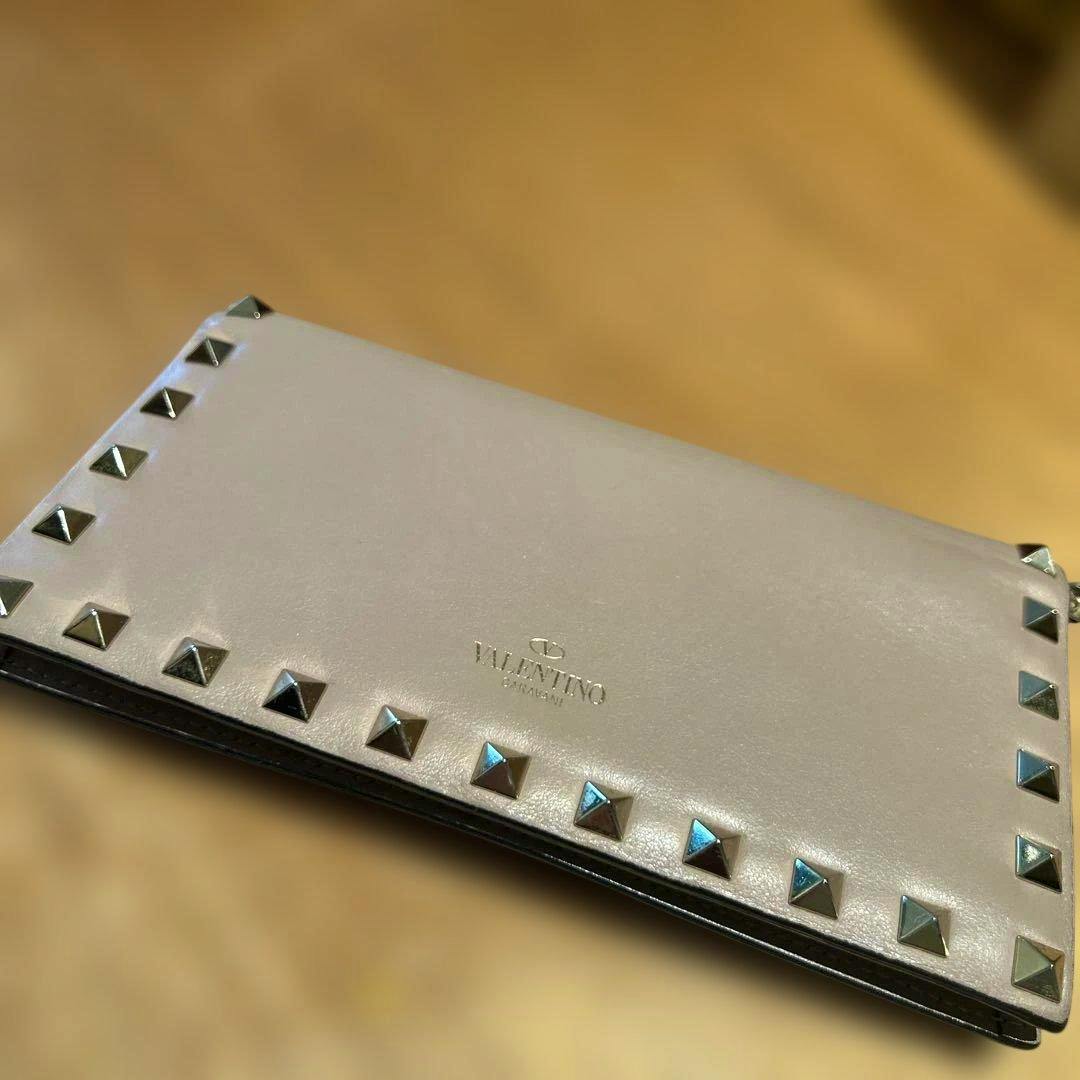 Light-colored Valentino Garavani long wallet with metal pyramid rockstuds along the edges and the Valentino logo embossed on the front, lying on a wooden surface.