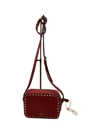 Red leather crossbody bag with square shape and metal stud trim hanging from its strap on a stand.