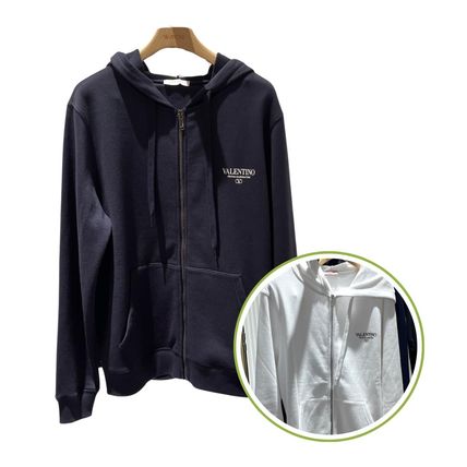 Dark hooded zip-up cotton sweatshirt with a small Valentino logo on the chest hanging on a wooden hanger, with a circular inset showing a white version.