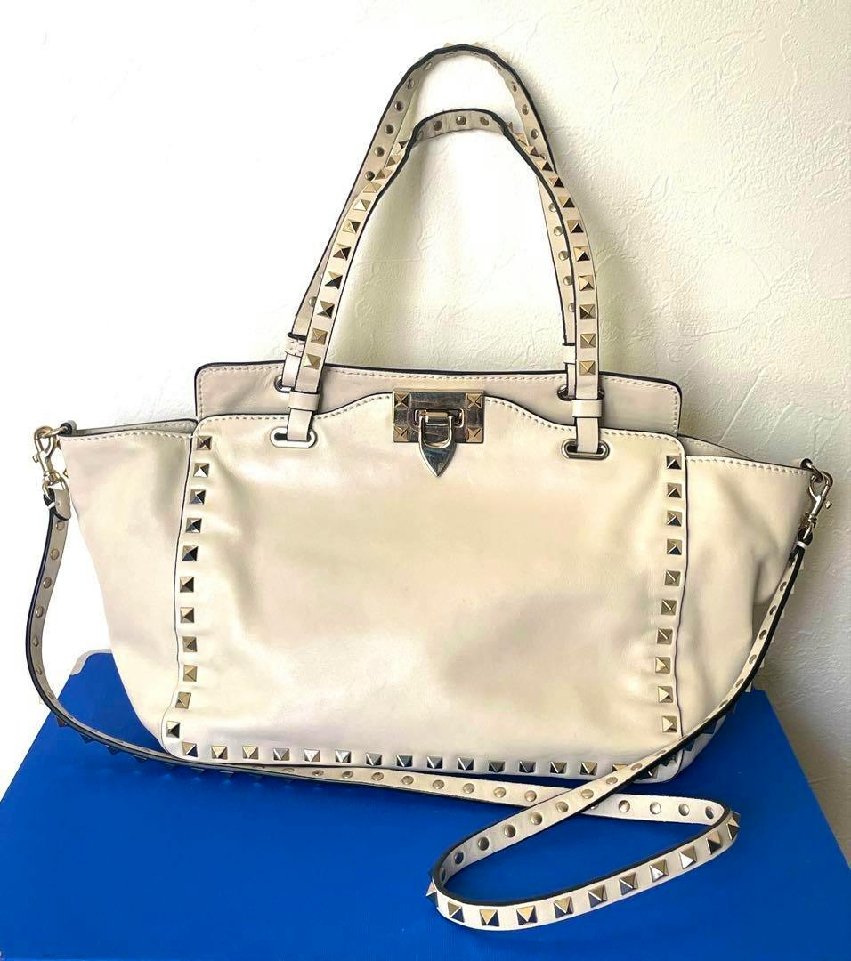 Front view of a Valentino Rockstud 2WAY bag in a light color with pyramid studs, top handles, a front clasp, and a detachable shoulder strap.