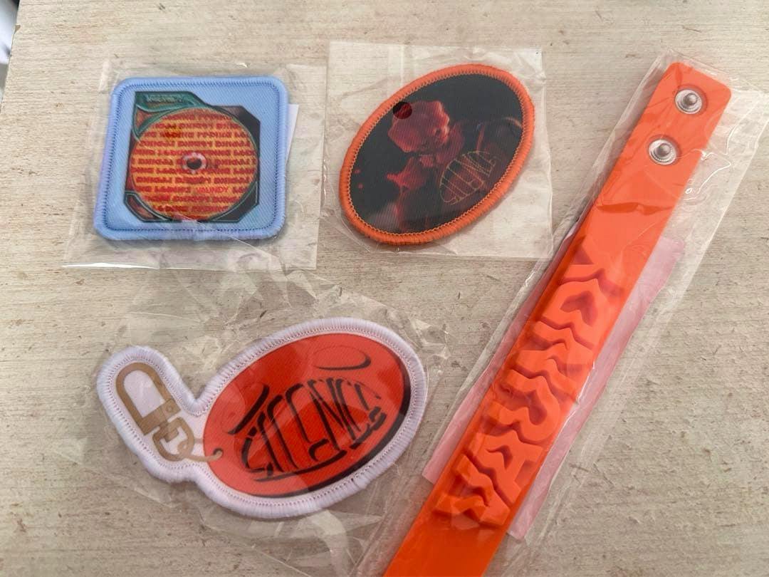 Three SILENCE-themed embroidered patches and an orange rubber wristband in plastic packaging laid on a light wood surface.