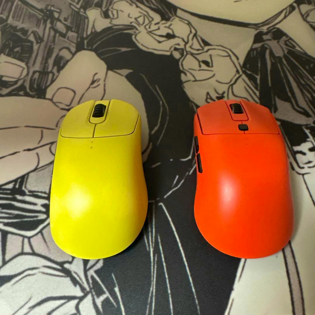 Top-down photo of two wireless computer mice side by side: a yellow mouse (left) and an orange mouse (right) on a patterned mat.
