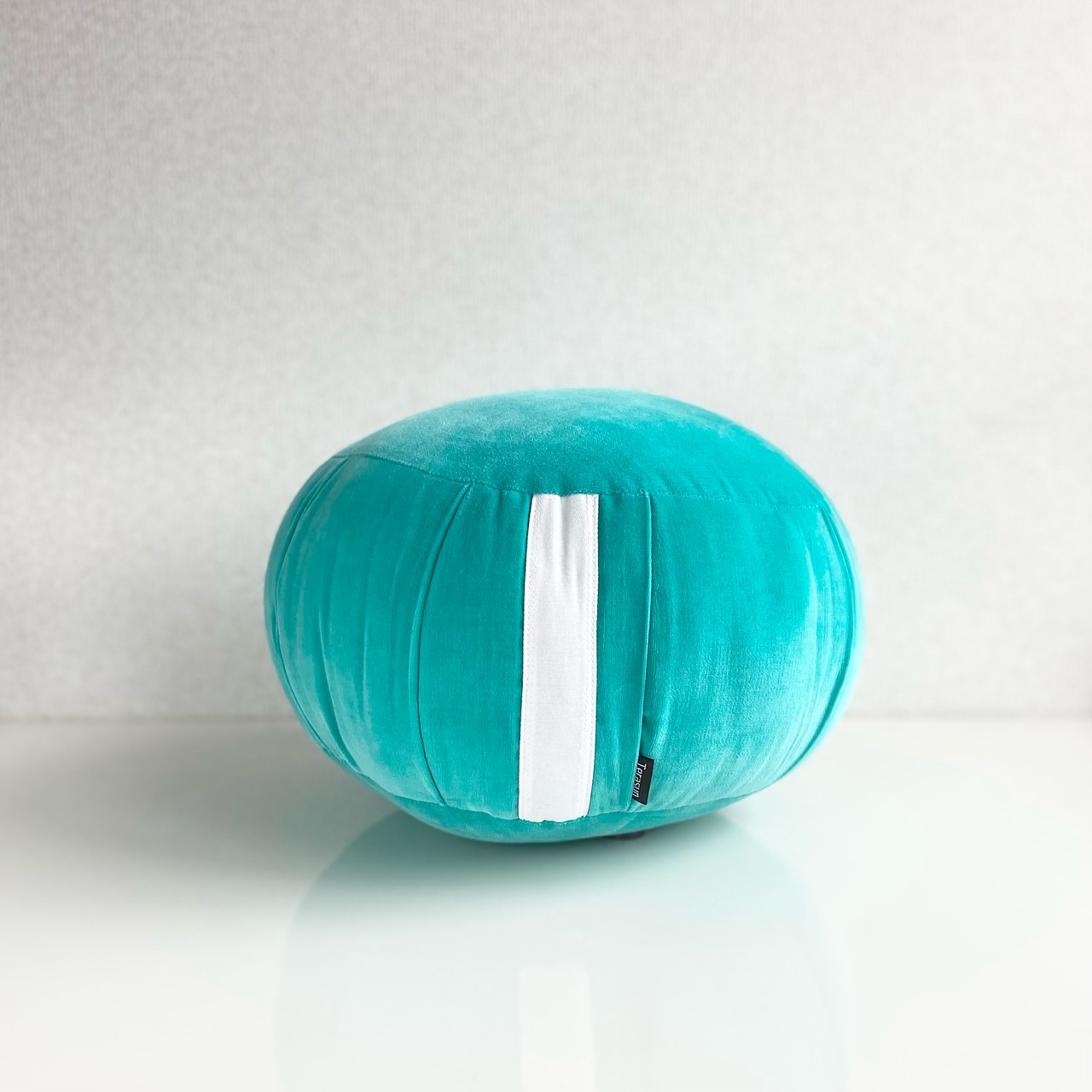 A teal velvet round sitting cushion with a white vertical band placed on a reflective surface against a pale background.