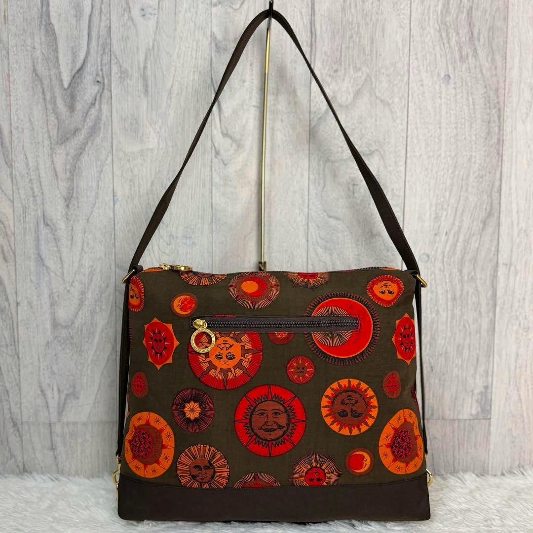 Versace sunburst canvas shoulder bag with brown background and orange-red sun motifs, front zipper pocket, hanging from its strap against a light wood backdrop.