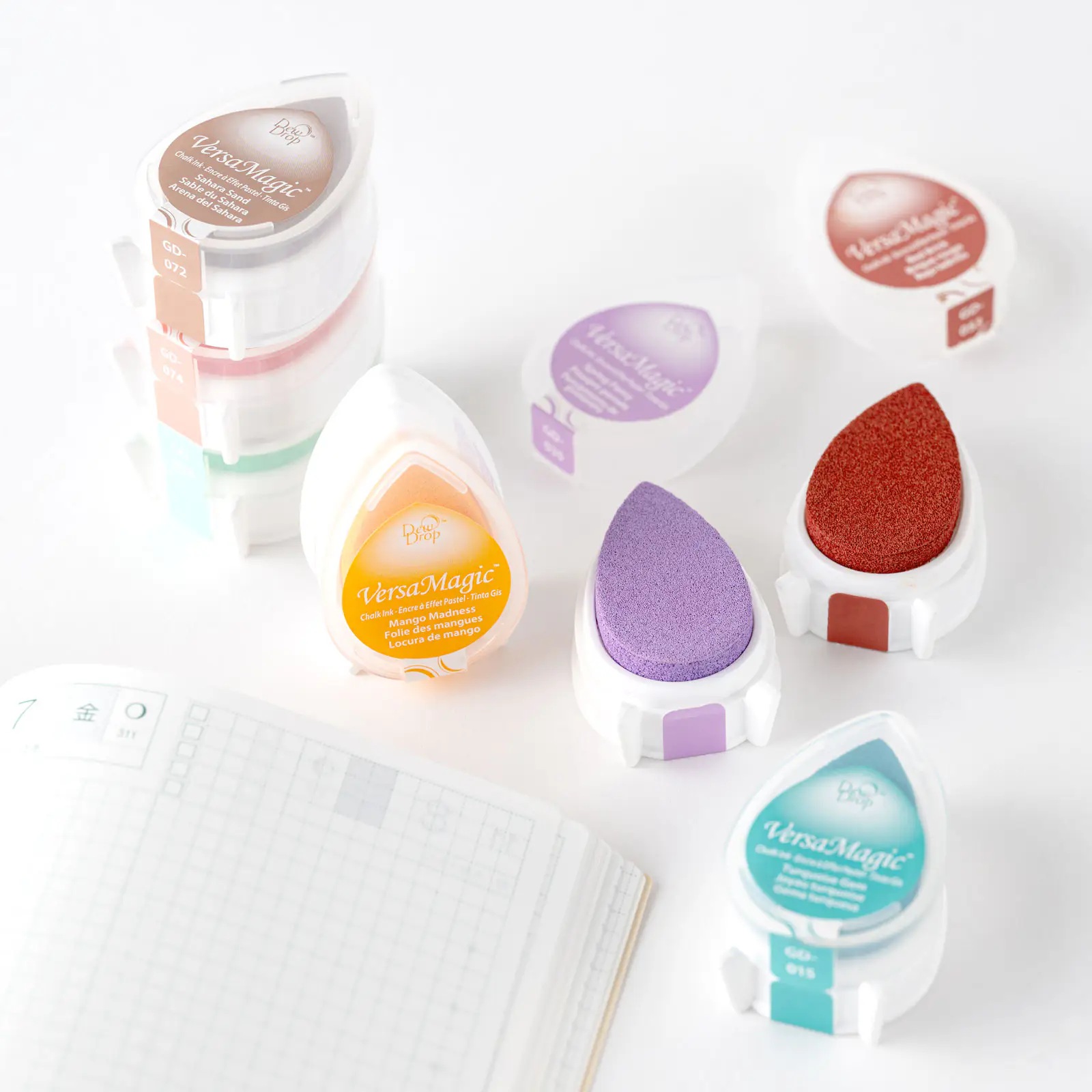 Several teardrop-shaped VersaMagic Dew Drop ink pads in different colors arranged on a white surface near an open notebook.