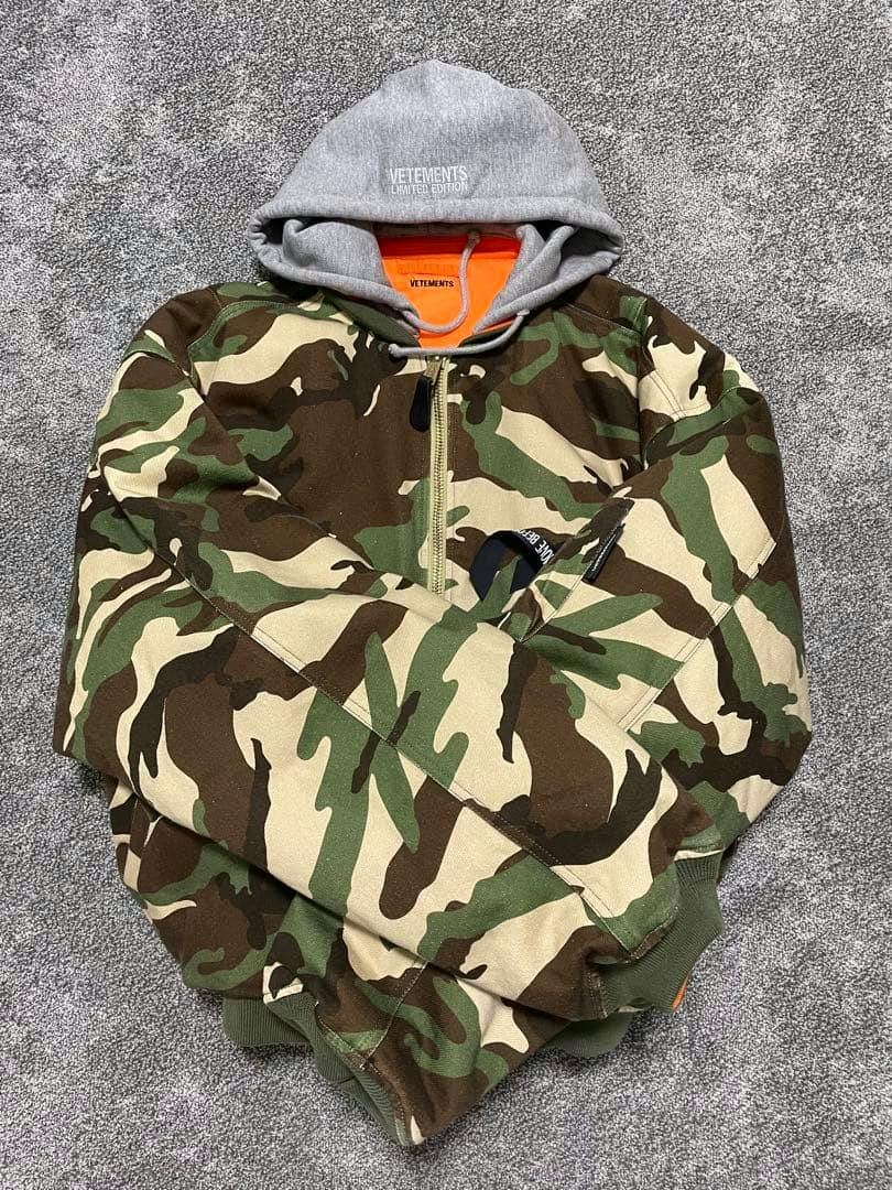 Camouflage VETEMENTS bomber jacket laid flat on a gray carpet, showing a gray hood with VETEMENTS label and an orange inner lining.
