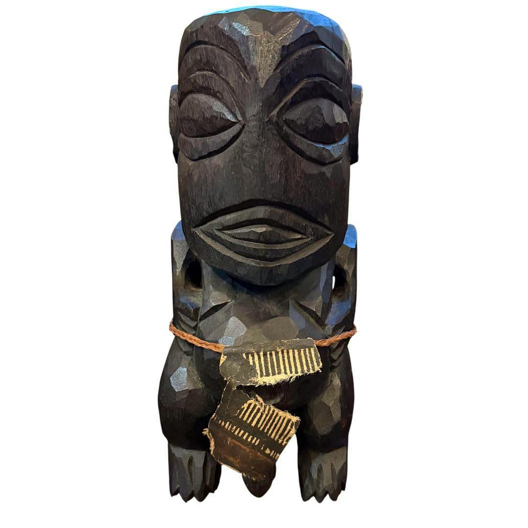 Front view of a carved wooden Tiki statue showing stylized facial features, a braided cord, and a small ornament at the waist.
