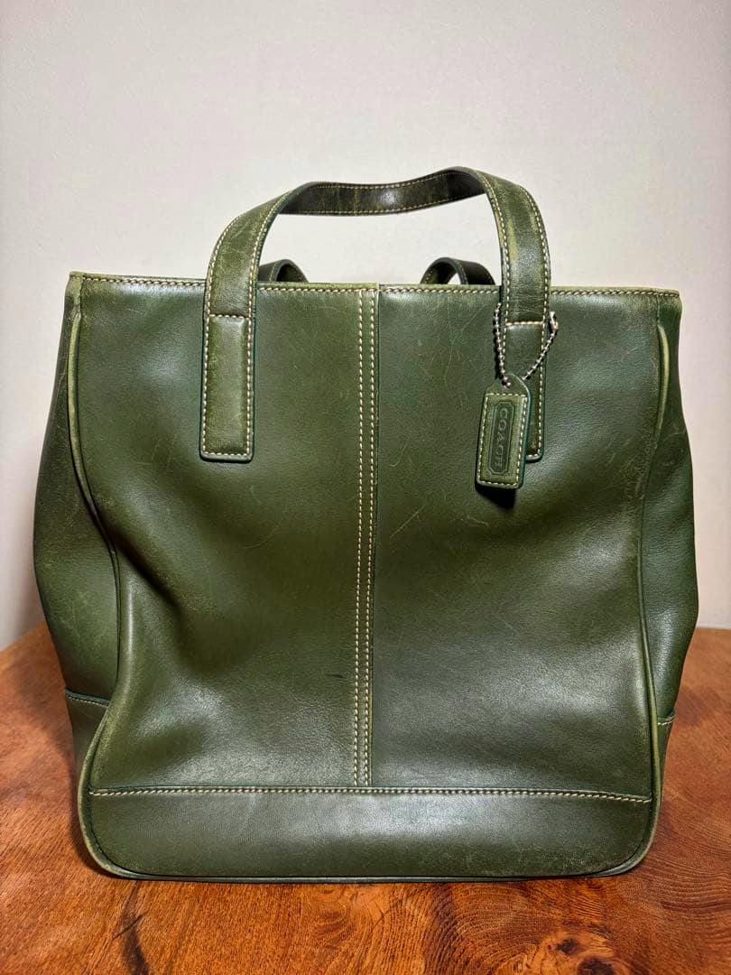 Green leather Coach tote handbag shown upright on a wooden surface from the front, with its hang tag visible.