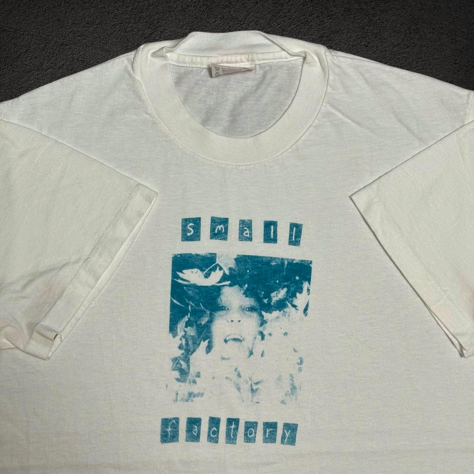 White T-shirt laid flat showing a teal printed photo-style band graphic with "small factory" text, ribbed neckline and short sleeves.
