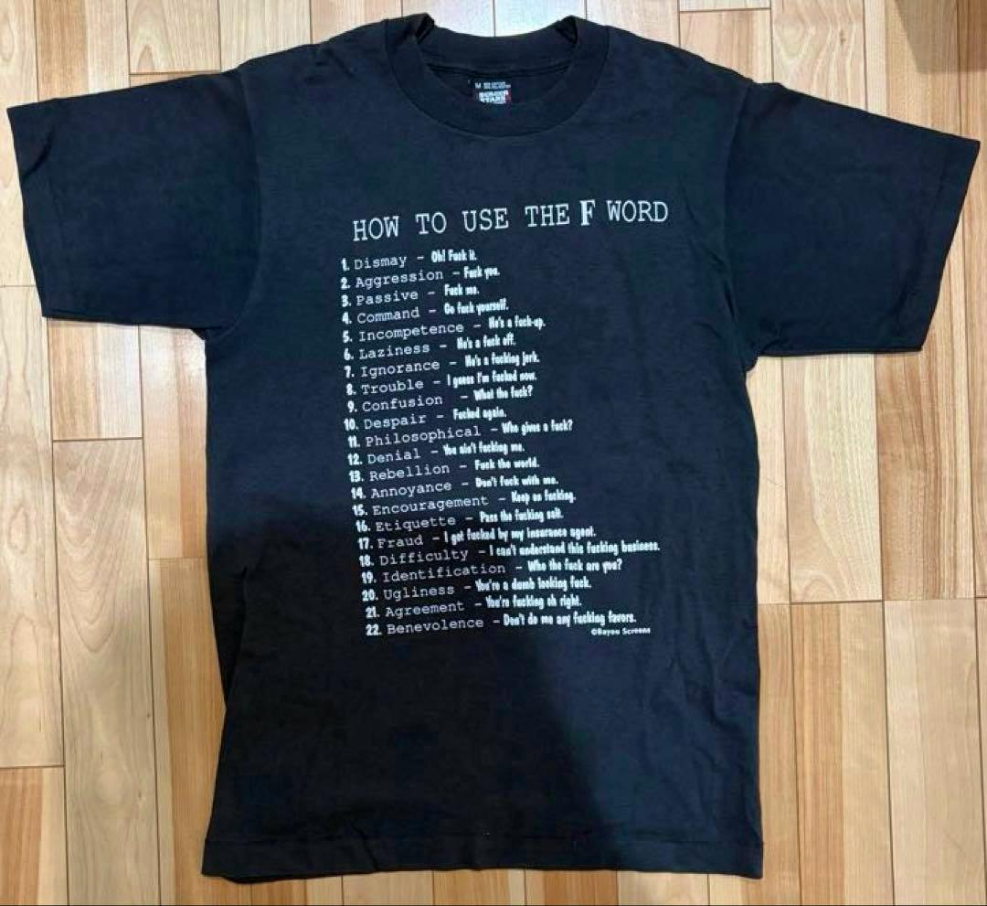 Front view of a black T-shirt laid flat showing the printed heading "HOW TO USE THE F WORD" and a numbered list of example phrases.