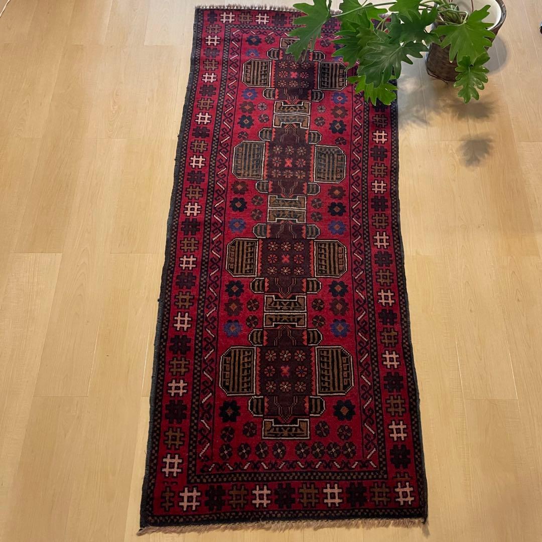 Red Baluchi tribal handwoven wool runner rug with geometric medallions laid on a light wood floor with a potted plant visible at the top right.