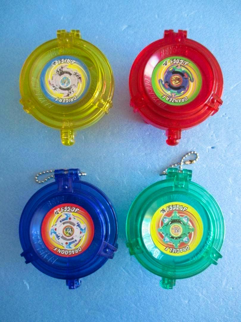 Four translucent round plastic Beyblade cases in yellow, red, blue, and green shown closed on a blue surface.