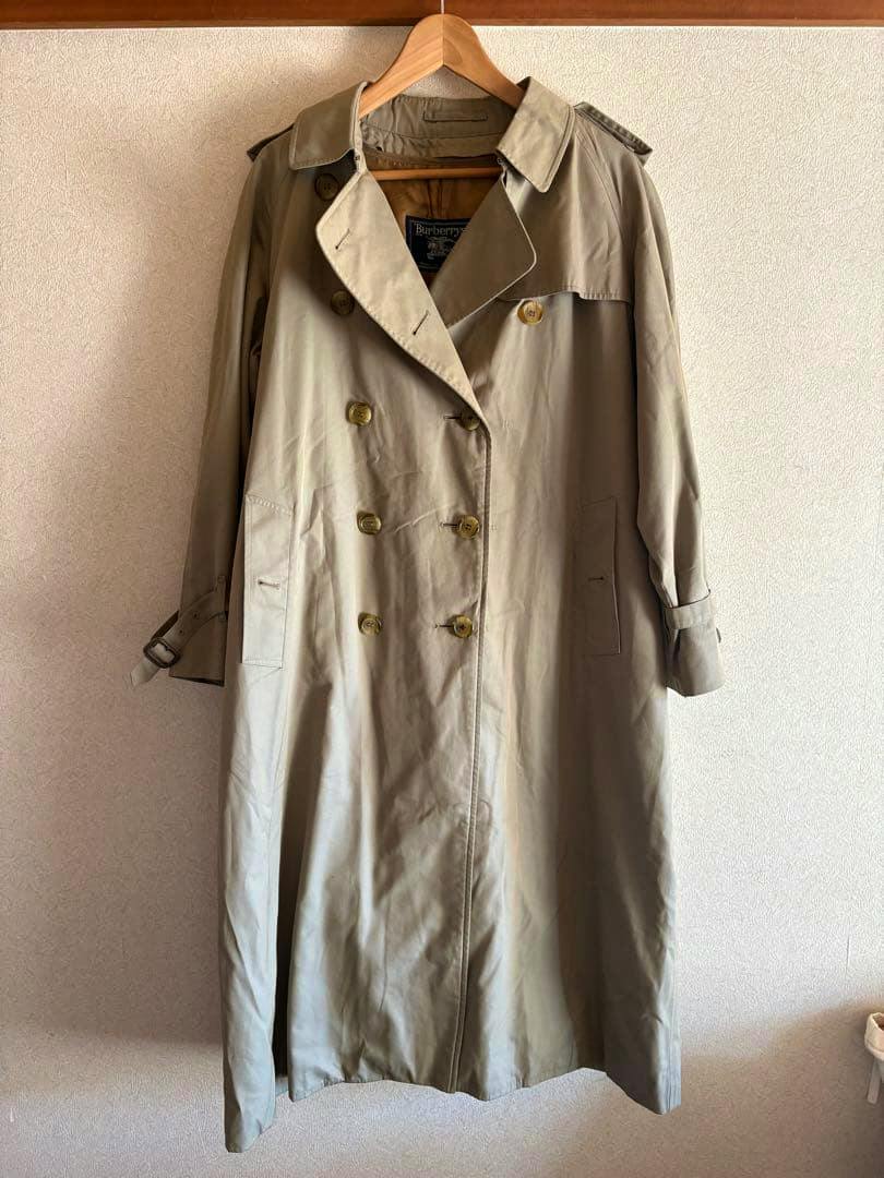 Beige women's Burberry trench coat hanging on a wooden hanger against a wall.