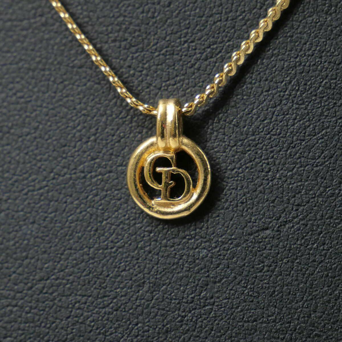 Close-up of a gold-color Christian Dior circular CD logo pendant on a chain against a dark textured background.