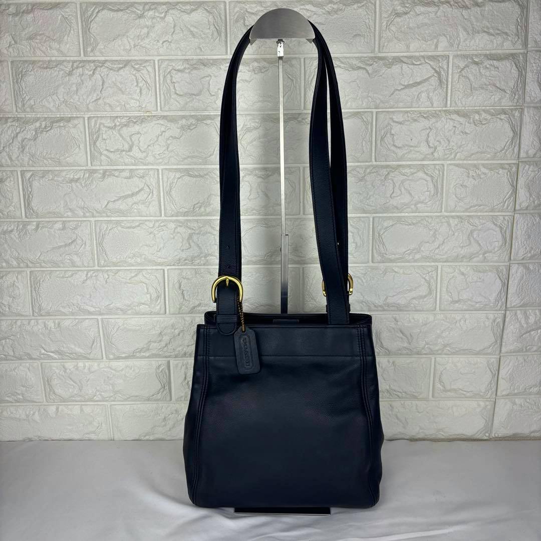 Front view of the leather Coach shoulder bag with long straps and a hanging leather tag against a white textured backdrop.