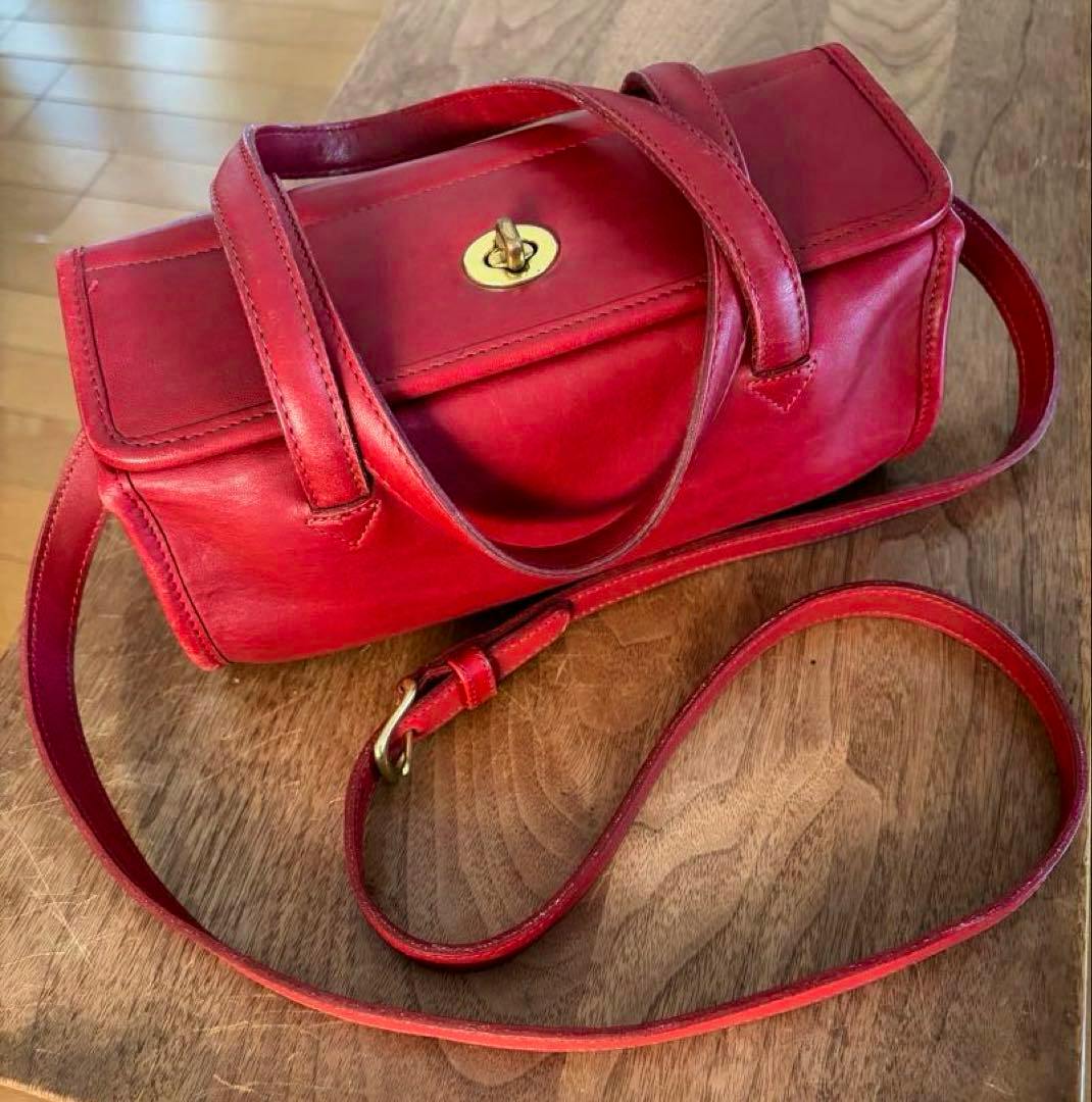 Red Coach cow leather 2-way shoulder bag with a gold turnlock and long strap photographed on a wooden surface.
