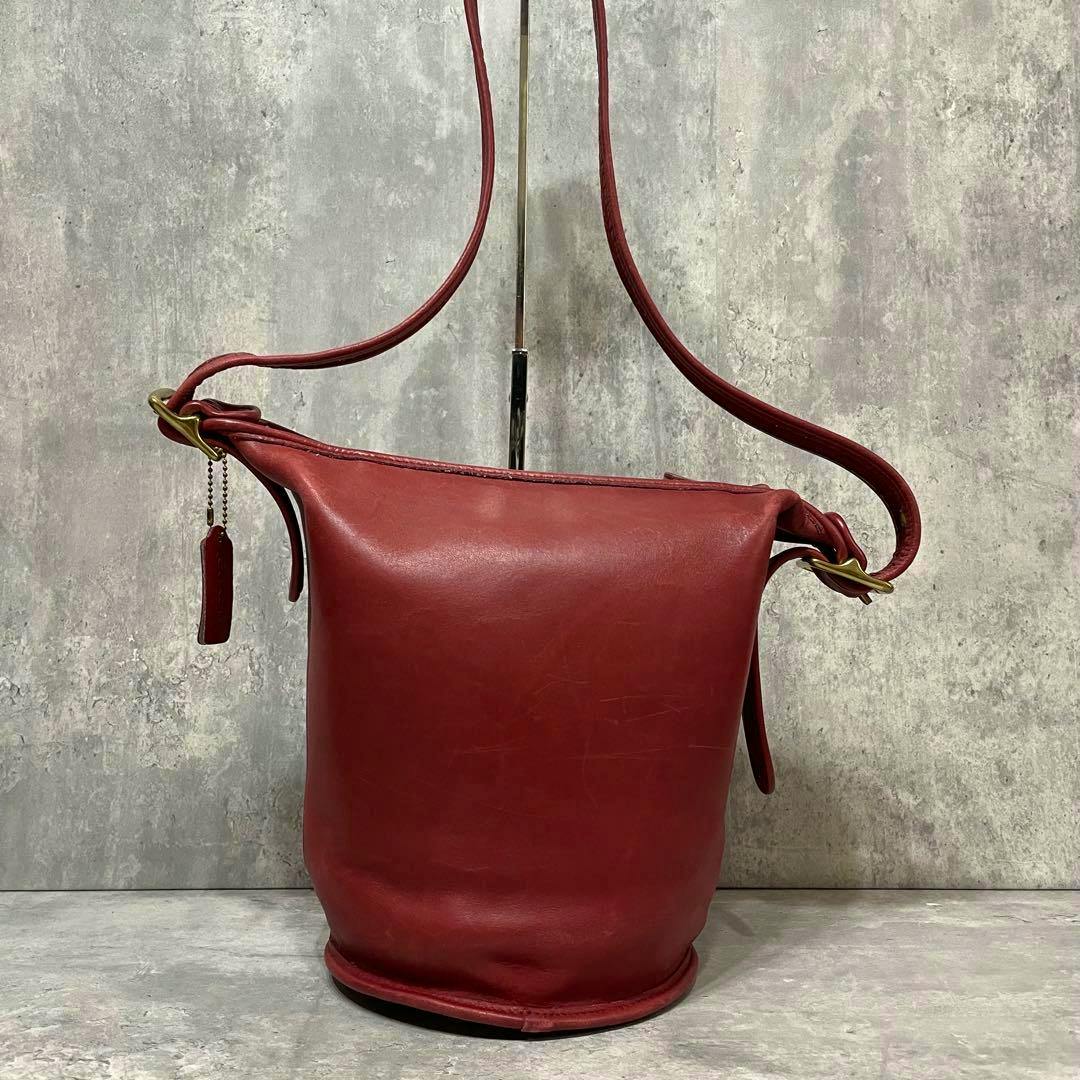 Front view of a red vintage Coach leather bucket bag with shoulder strap and a small leather hangtag against a gray textured background.