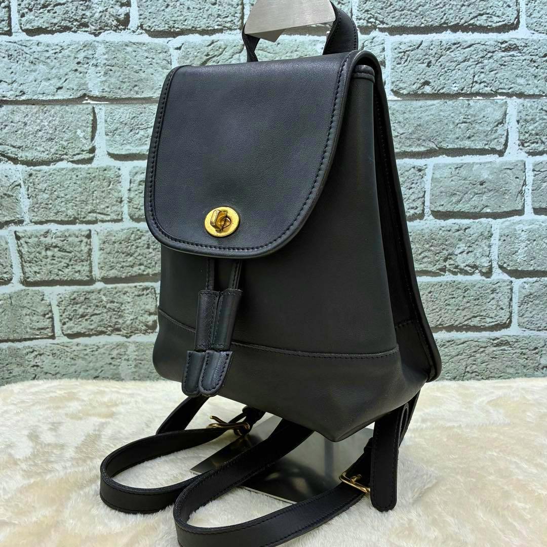 Black leather mini backpack with a gold turnlock flap and two shoulder straps shown against a brick wall.