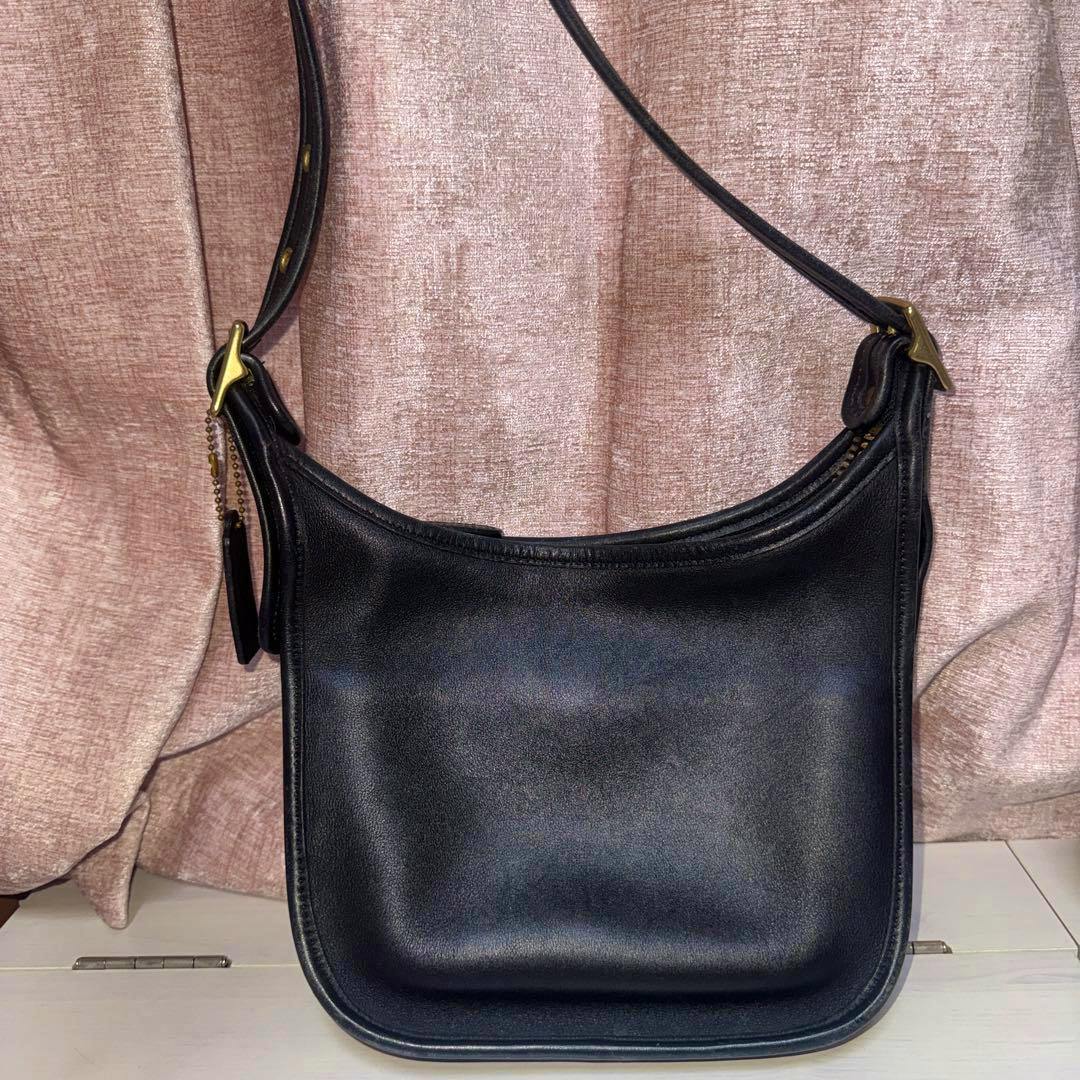 Front view of a black leather Coach shoulder bag with an adjustable strap and brass hardware against a pink curtain backdrop.