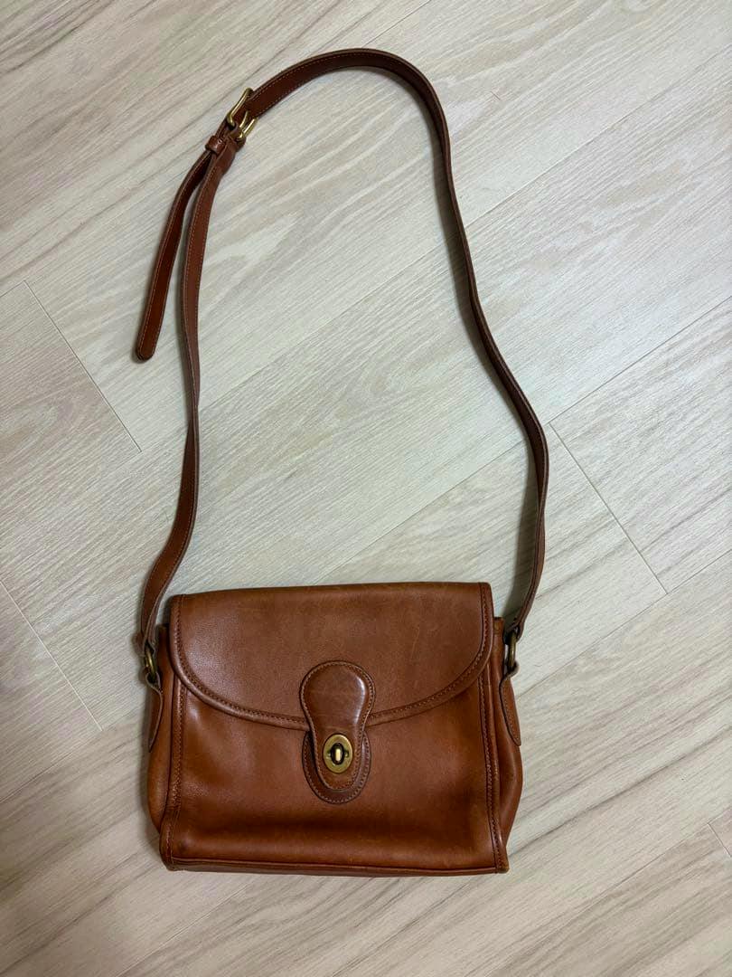 Vintage COACH brown leather shoulder bag with a flap and turn-lock closure laid flat on a light wood floor.