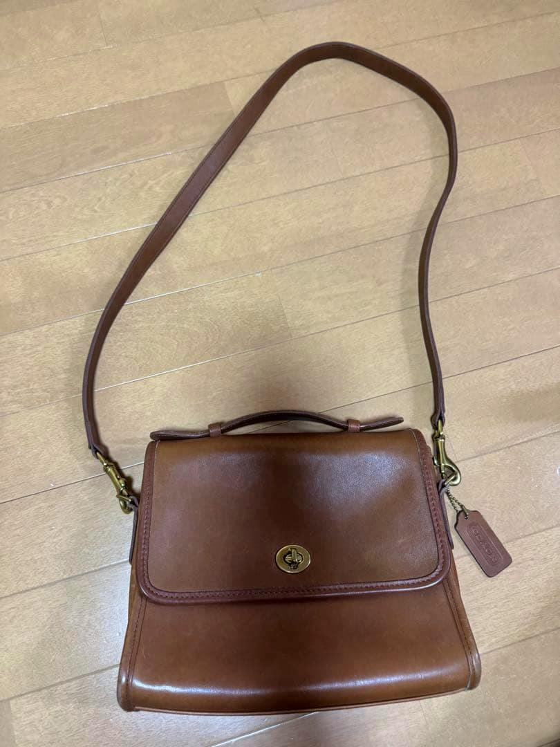 Brown leather Coach bag with flap and turn-lock, top handle and shoulder strap on a wooden floor.