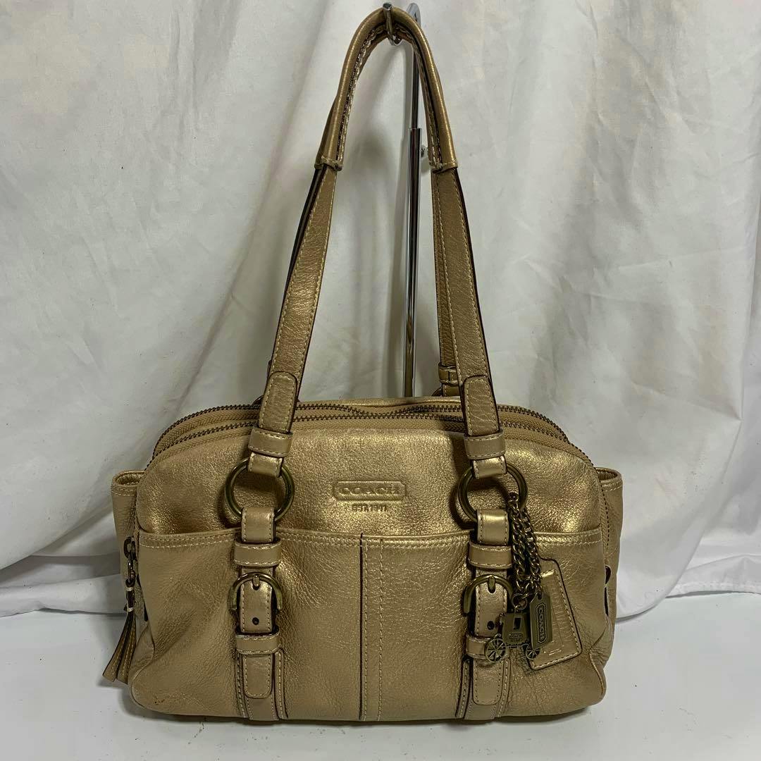Front view of a gold leather Coach handbag with double handles, front pockets, buckles, and hanging metal Coach charms.