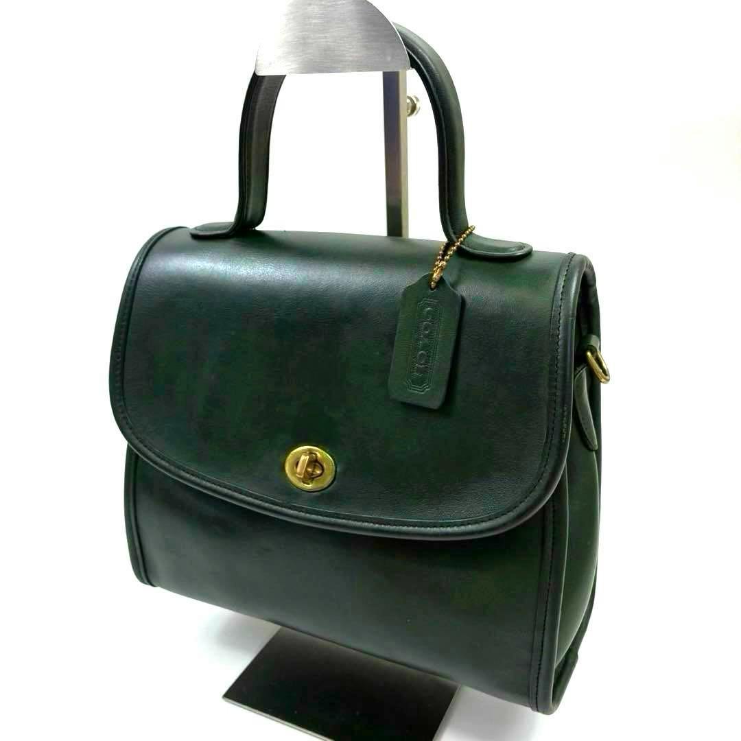 Green Coach cow leather handbag with top handle, gold turn-lock closure, and Coach hang tag on a display stand.