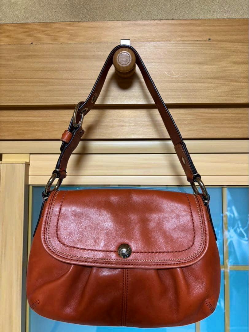 Front view of a brown leather Coach handbag with a flap closure and single shoulder strap hanging from a wooden peg.