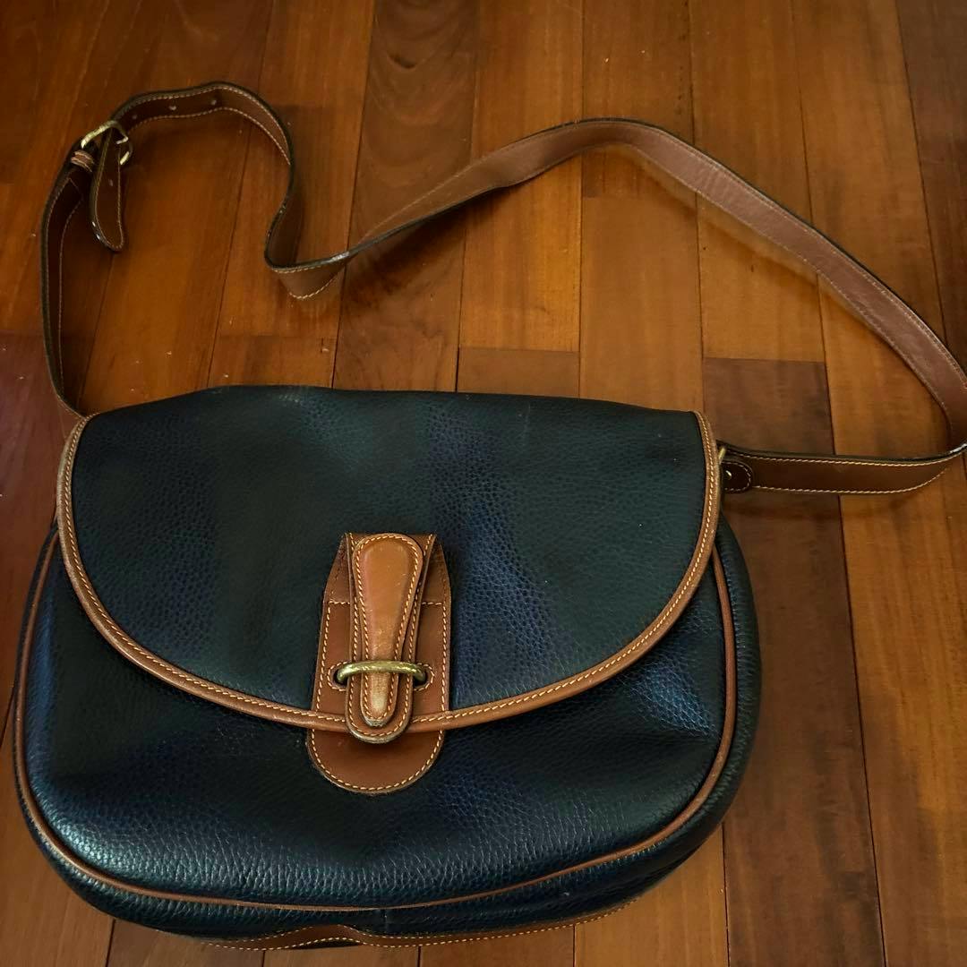 Old Coach leather handbag lying on a wooden floor, showing a pebbled dark body with brown leather trim, flap, and shoulder strap.
