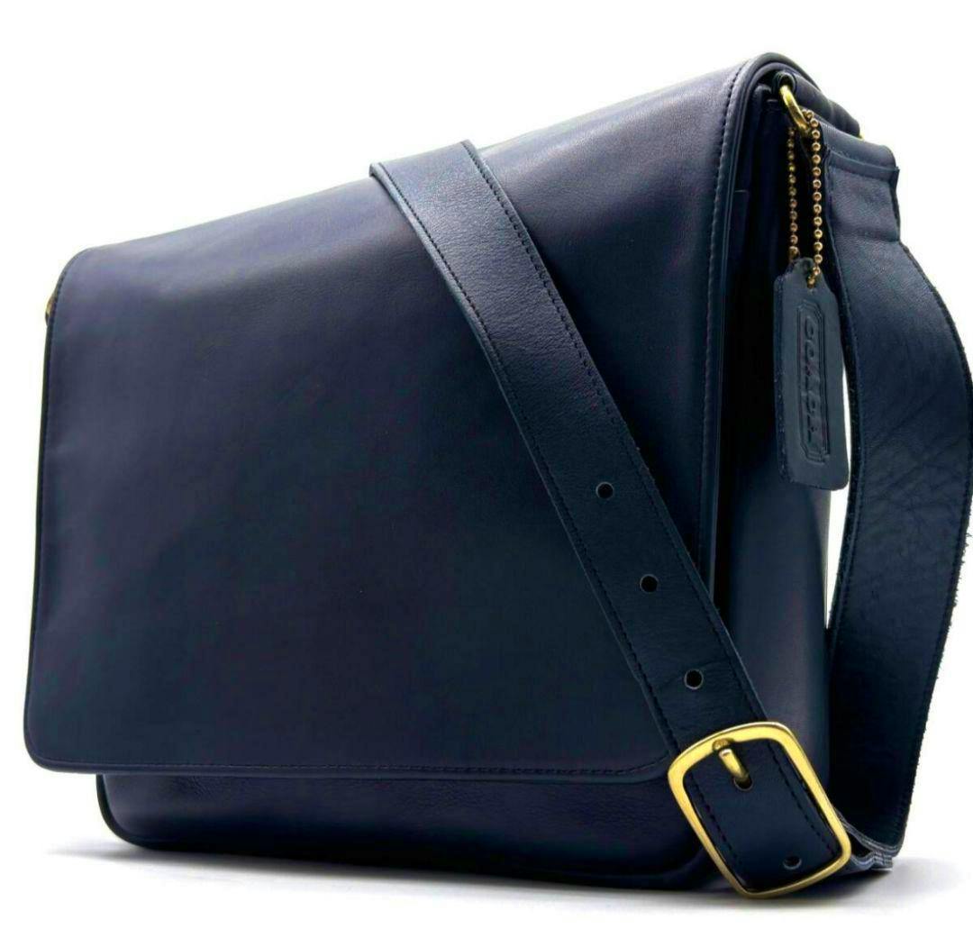 Three-quarter view of the Old Coach cow leather shoulder bag showing the flap, adjustable strap, leather hang tag, and gold-tone buckle.