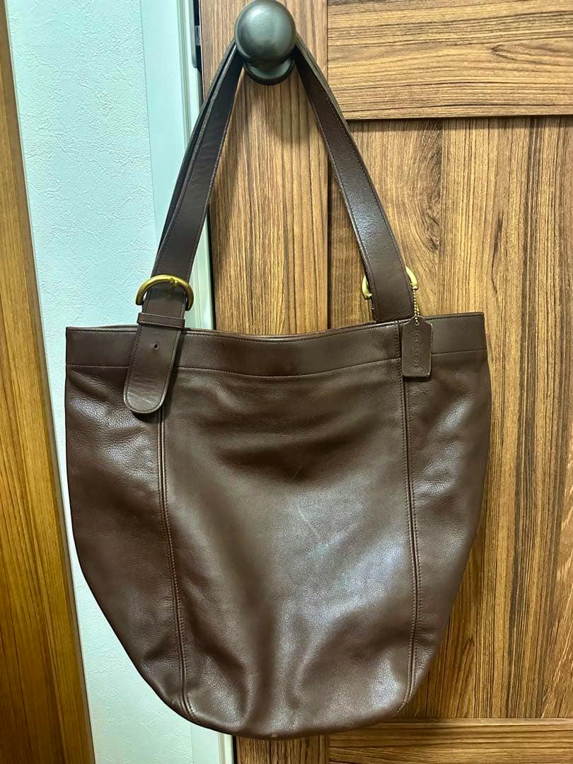 Brown COACH leather tote bag hanging on a door knob, front view showing straps and body.