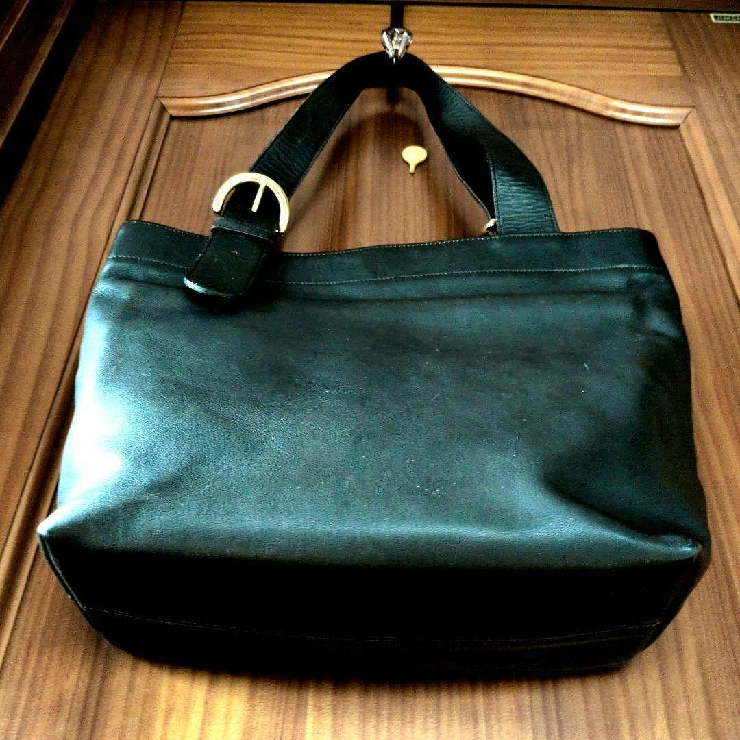 Black leather tote bag hanging on a wooden door, front view showing straps and a metal buckle.
