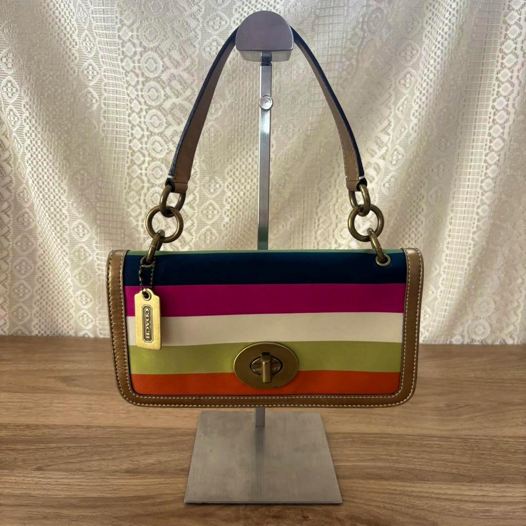 Front view of a vintage Coach nylon shoulder bag with horizontal colored stripes, leather trim, a turn-lock closure, and a hanging Coach tag.
