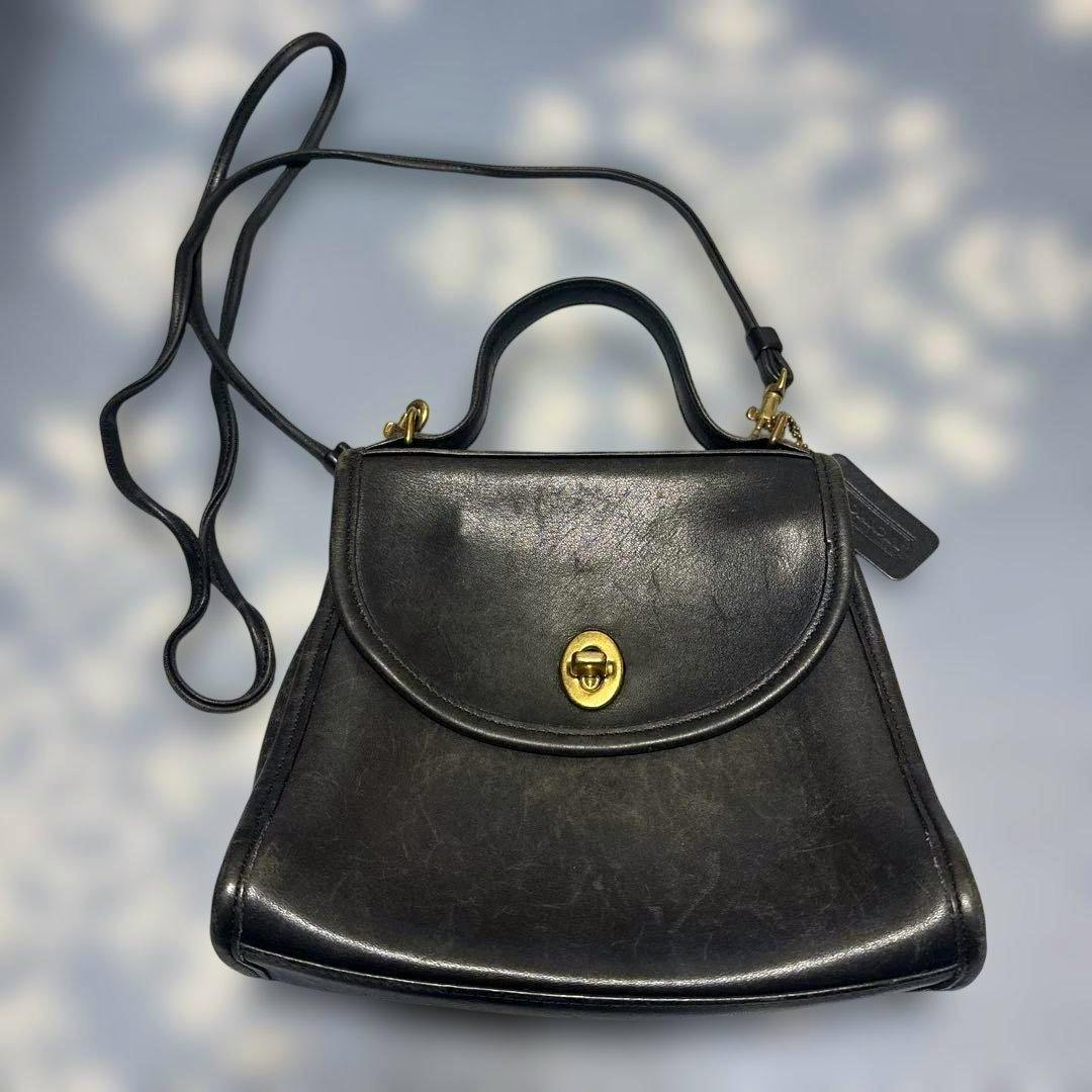 Black Old Coach cow leather shoulder bag with a top handle, long strap, and gold turn-lock closure shown laid flat.