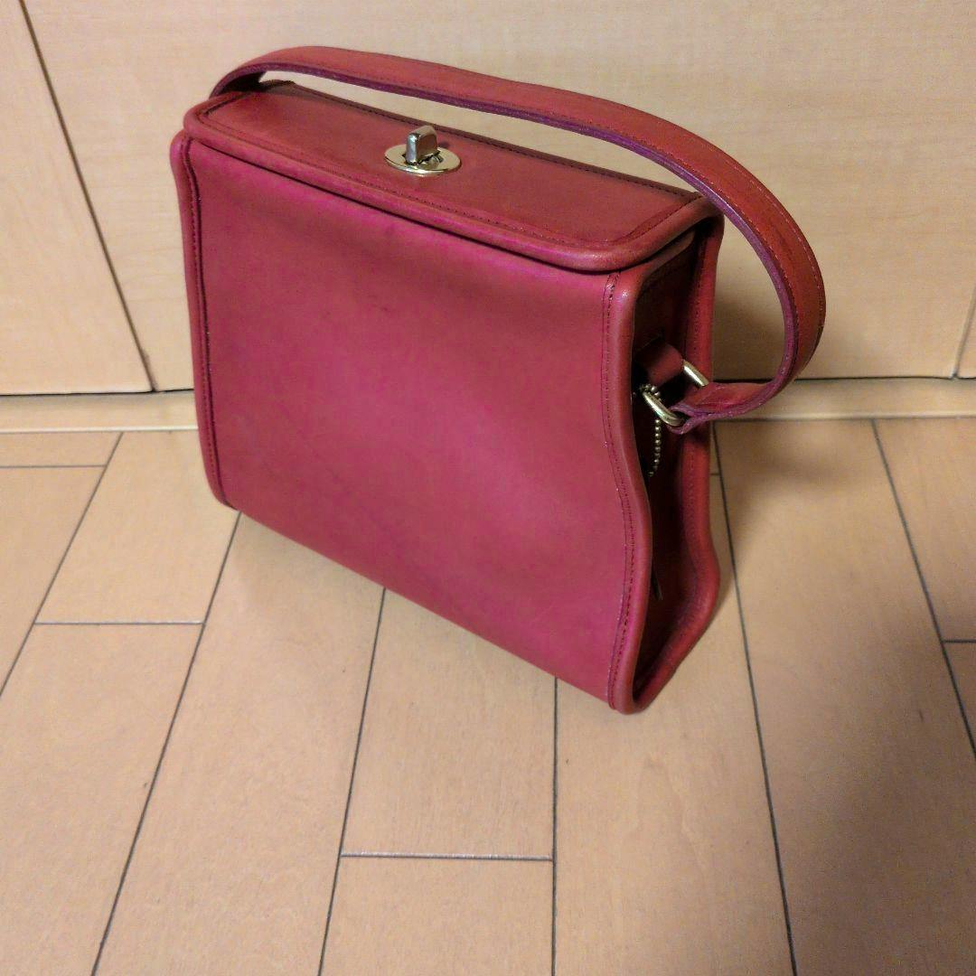 Red leather handbag on a wooden floor shown at an angled front-side view with a top turn-lock and single strap.