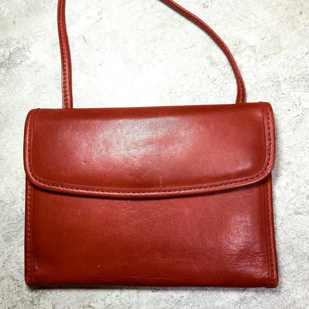 Front view of a red leather shoulder bag pouch/wallet with a flap and thin shoulder strap on a light textured surface.