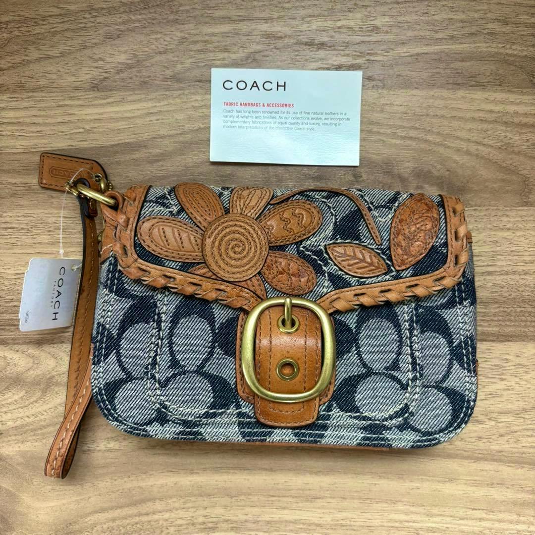Front view of a Coach signature denim pouch with leather flower appliqué, braided trim, brass buckle, and a leather wrist strap on a wooden surface.