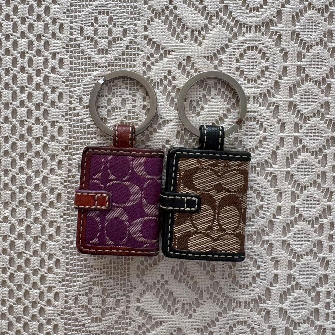 Two vintage Coach signature photo keychain charms, one purple and one brown, displayed on a lace background.