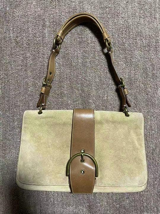 Front view of a beige suede Coach shoulder handbag with a brown leather center strap and metal buckle on a carpet.