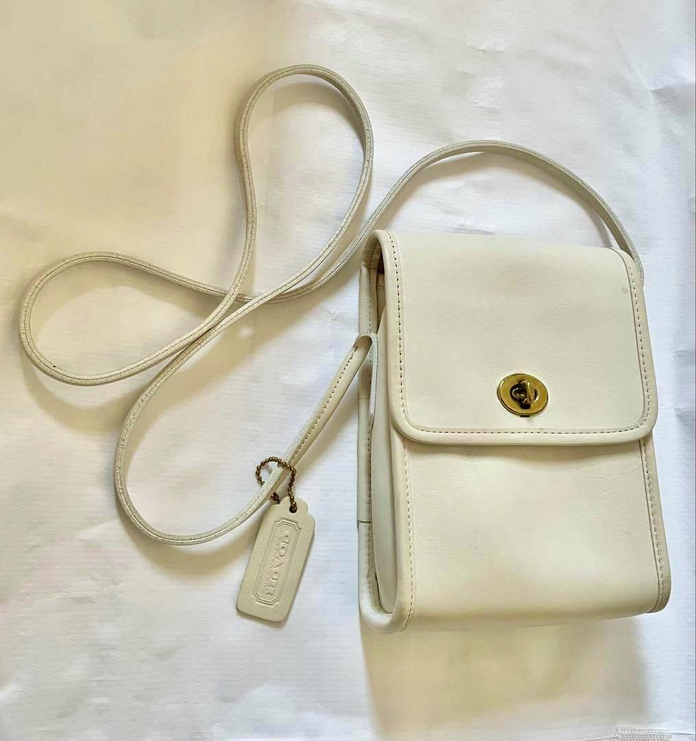 Ivory leather Coach shoulder bag with a turn-lock flap and hanging Coach tag on a long strap, photographed on a white background.