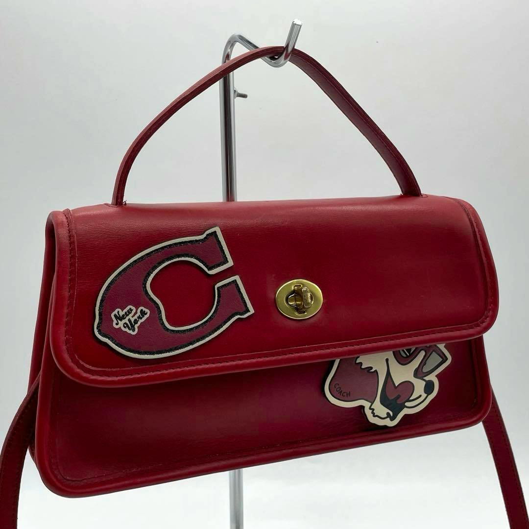 Red Coach turnlock shoulder bag with top handle, gold turnlock, and decorative patches displayed on a stand.