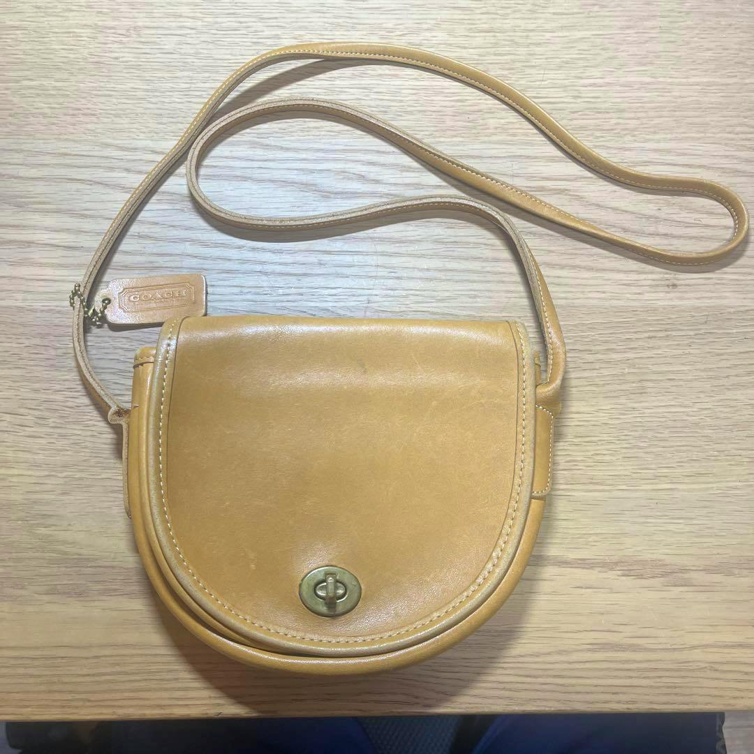 Camel leather vintage Coach mini shoulder bag in a half-moon shape with a turn-lock closure and strap, shown on a wooden surface.