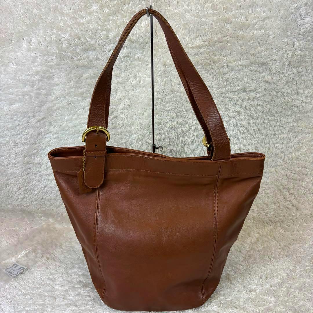 Brown Coach glove-tan leather large tote bag displayed upright with shoulder straps on a light textured backdrop.