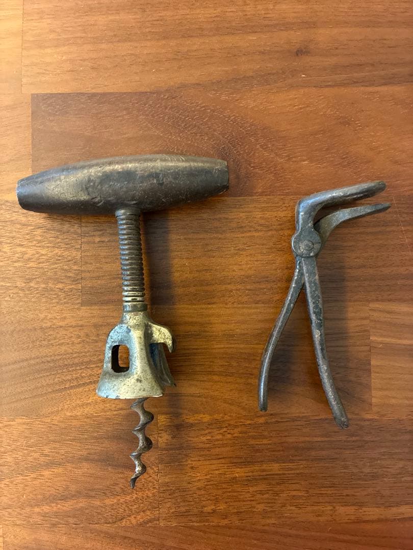 Two vintage metal bottle-opening tools—a T-handle corkscrew and a plier-style opener—laid on a wooden surface.