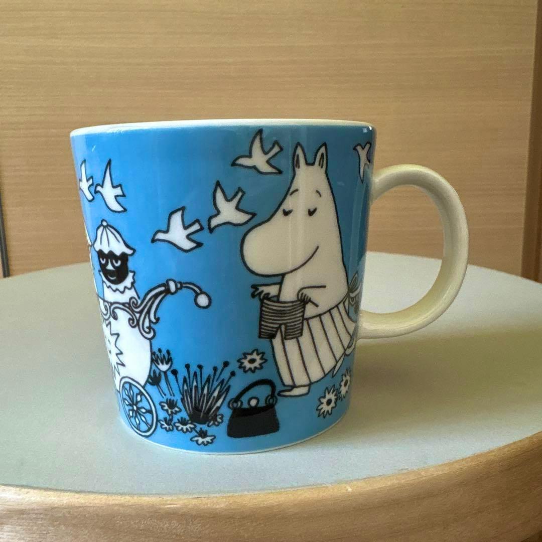 Photo of an ARABIA Moomin mug with a blue background showing the Moomin character, birds, and floral motifs on a table.