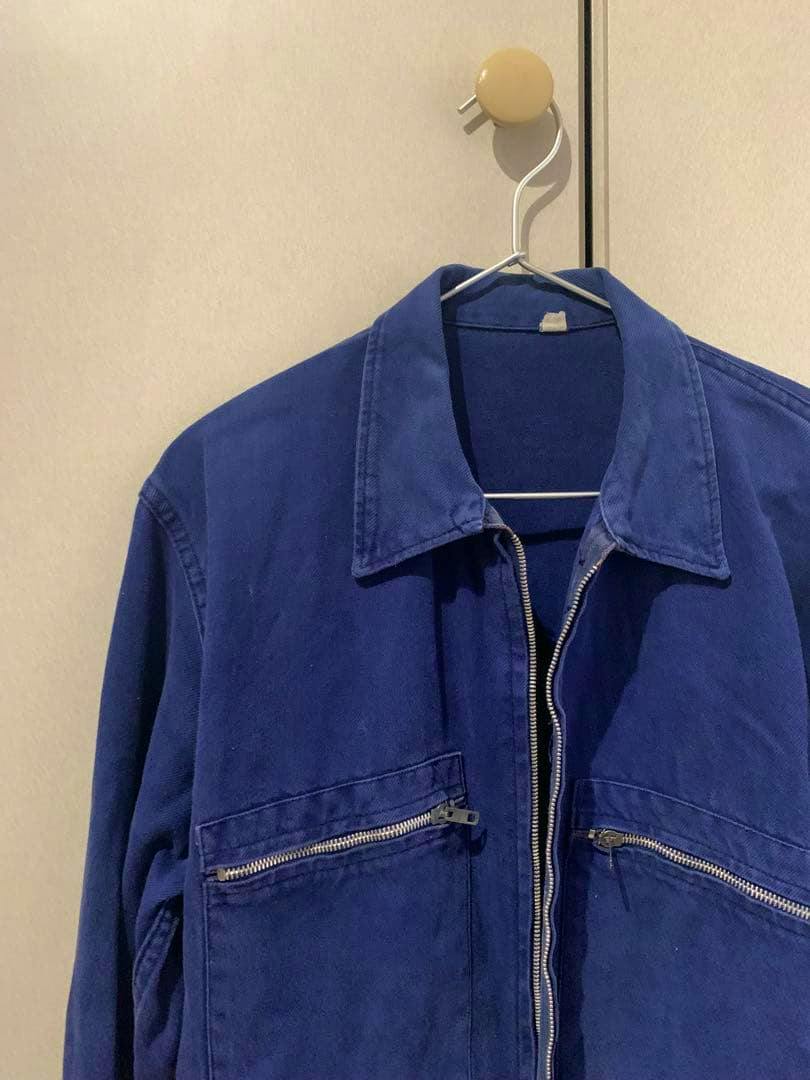 Blue men's cotton work jacket hanging on a hanger showing the front with collar, central zipper, and two zippered chest pockets.