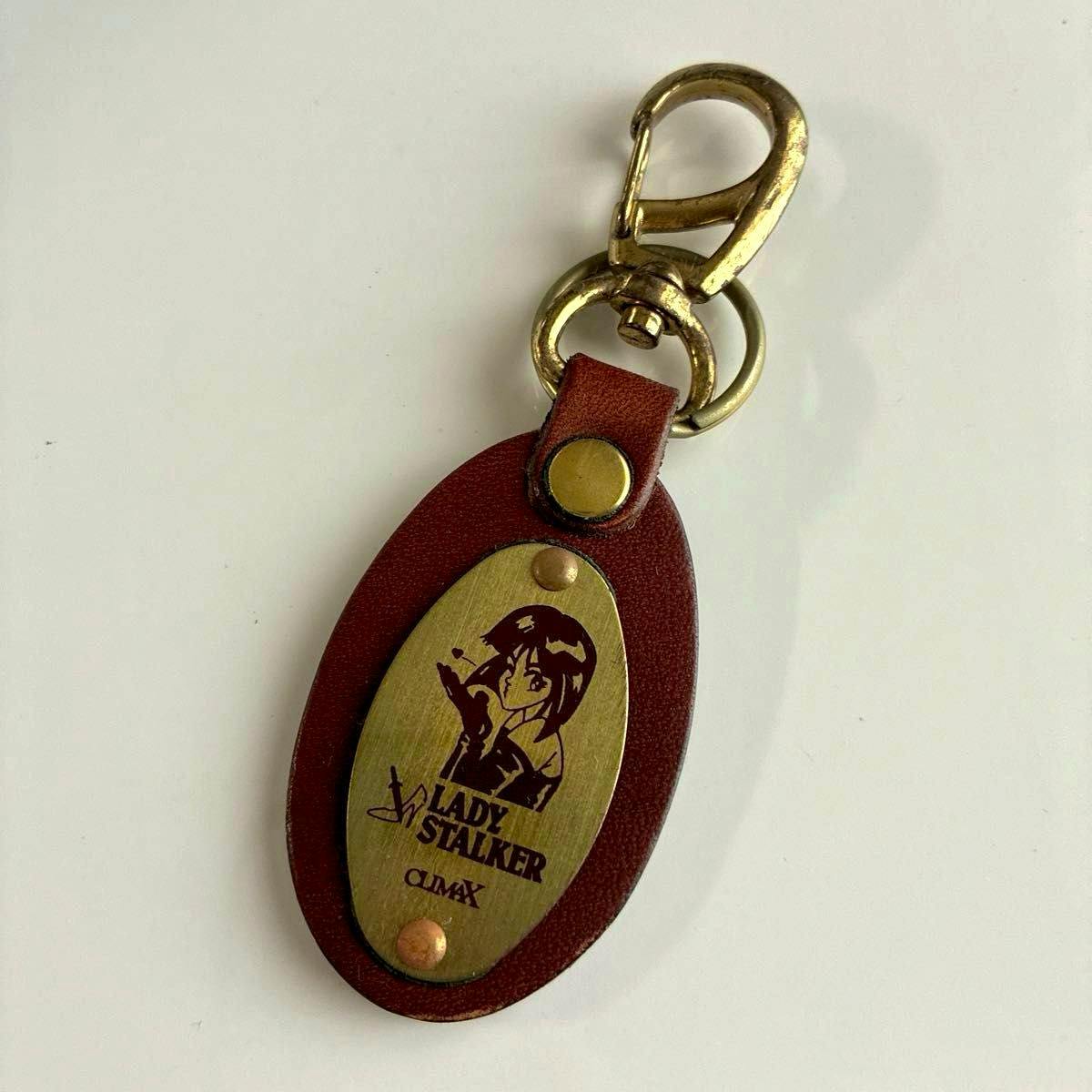 Brown cow-leather oval keychain with a brass oval plate printed with a Lady Stalker character and a brass clasp.