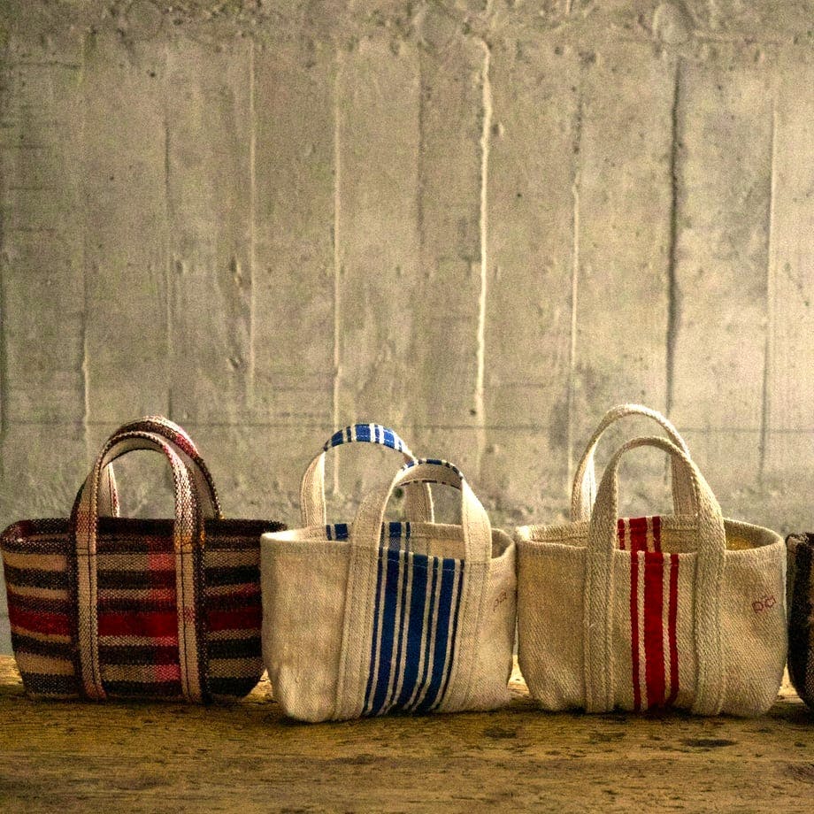 Three small vintage linen canvas tote bags with striped patterns arranged side by side on a wooden surface against a concrete wall background.
