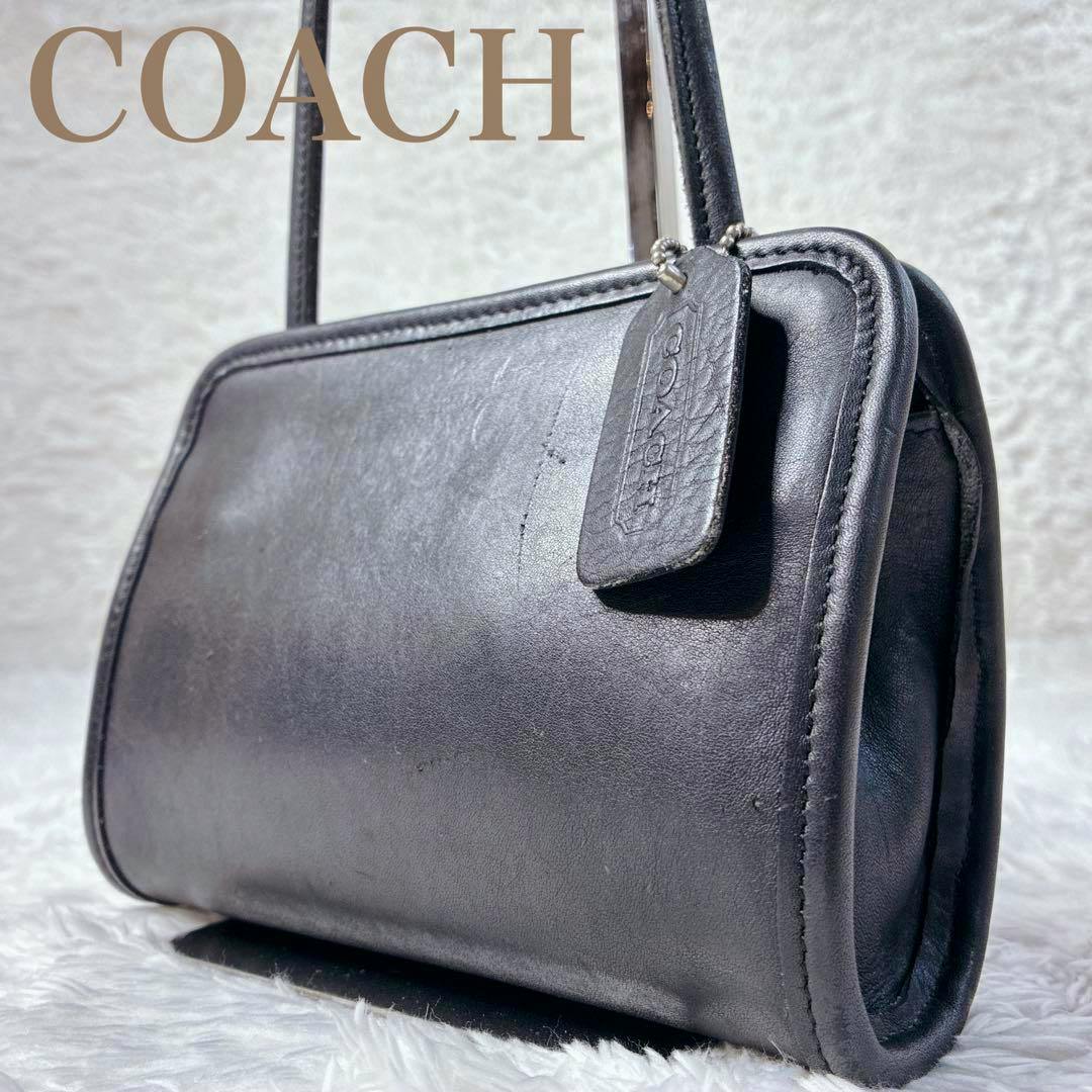 Black all-leather vintage Coach shoulder bag shown at an angled view with an attached leather hang tag and narrow shoulder straps.